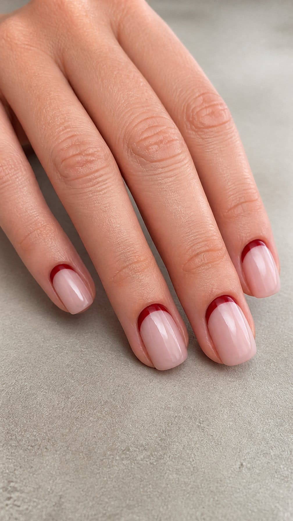 A hand with glossy nude nails featuring deep red reverse French tips near the cuticles.
