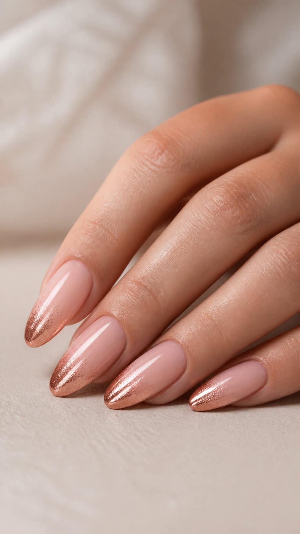 A hand with almond-shaped nails featuring a nude-to-rose-gold ombre manicure.