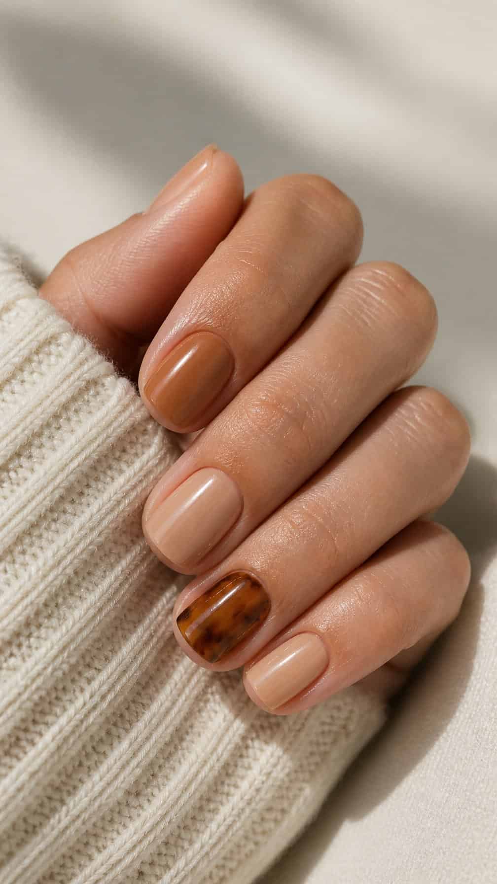 A close-up of a hand with short manicured nails featuring glossy neutral polish and tortoiseshell accent nails.