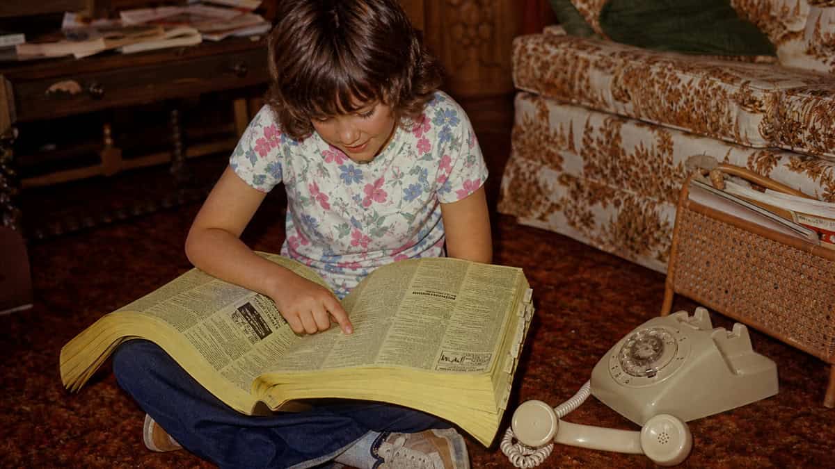14 Things Every Kid Who Used a Rotary Phone Remembers 6 Child balancing a heavy telephone directory beside a rotary phone in the 1970s