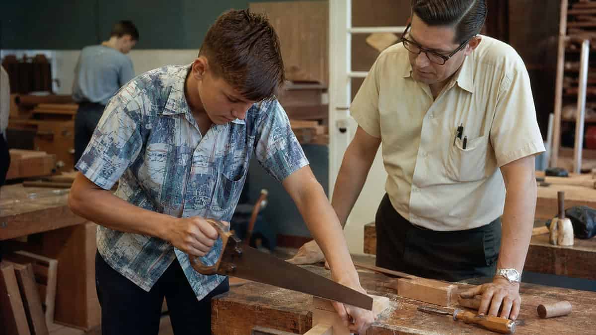 Teenager sawing a board under teacher supervision in a 1960s shop class