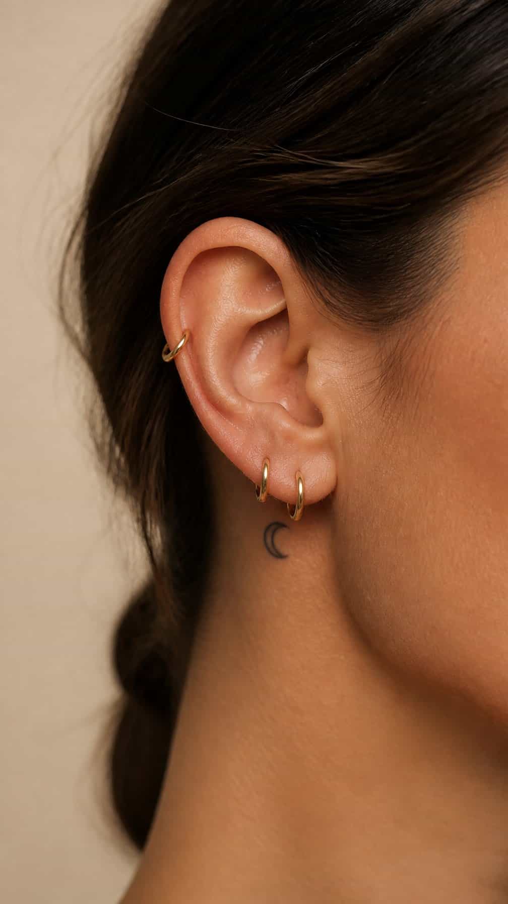A close-up of an ear with tiny gold hoop earrings and a small crescent-moon tattoo.