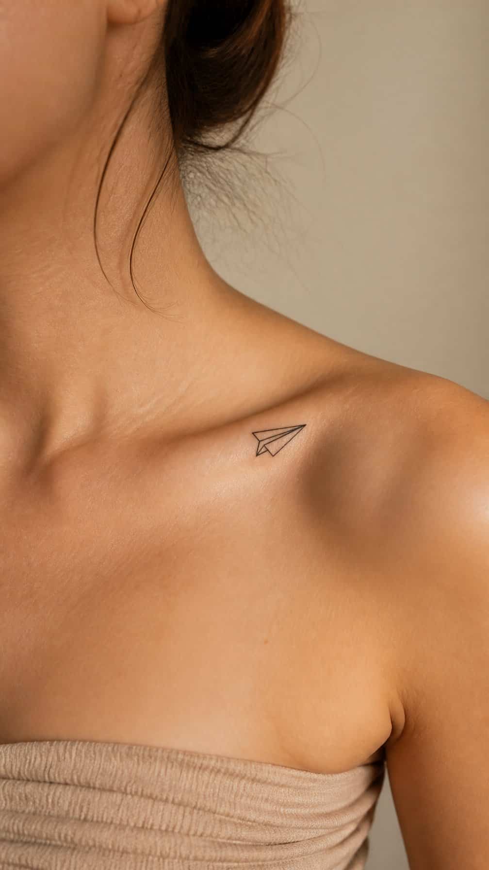 A small paper airplane tattoo angled toward the shoulder beneath a person’s collarbone.
