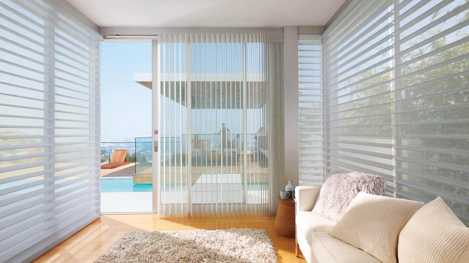 Bright, airy living space featuring expansive windows, vertical blinds, cozy sofa, and a view of the balcony and outdoor pool area.