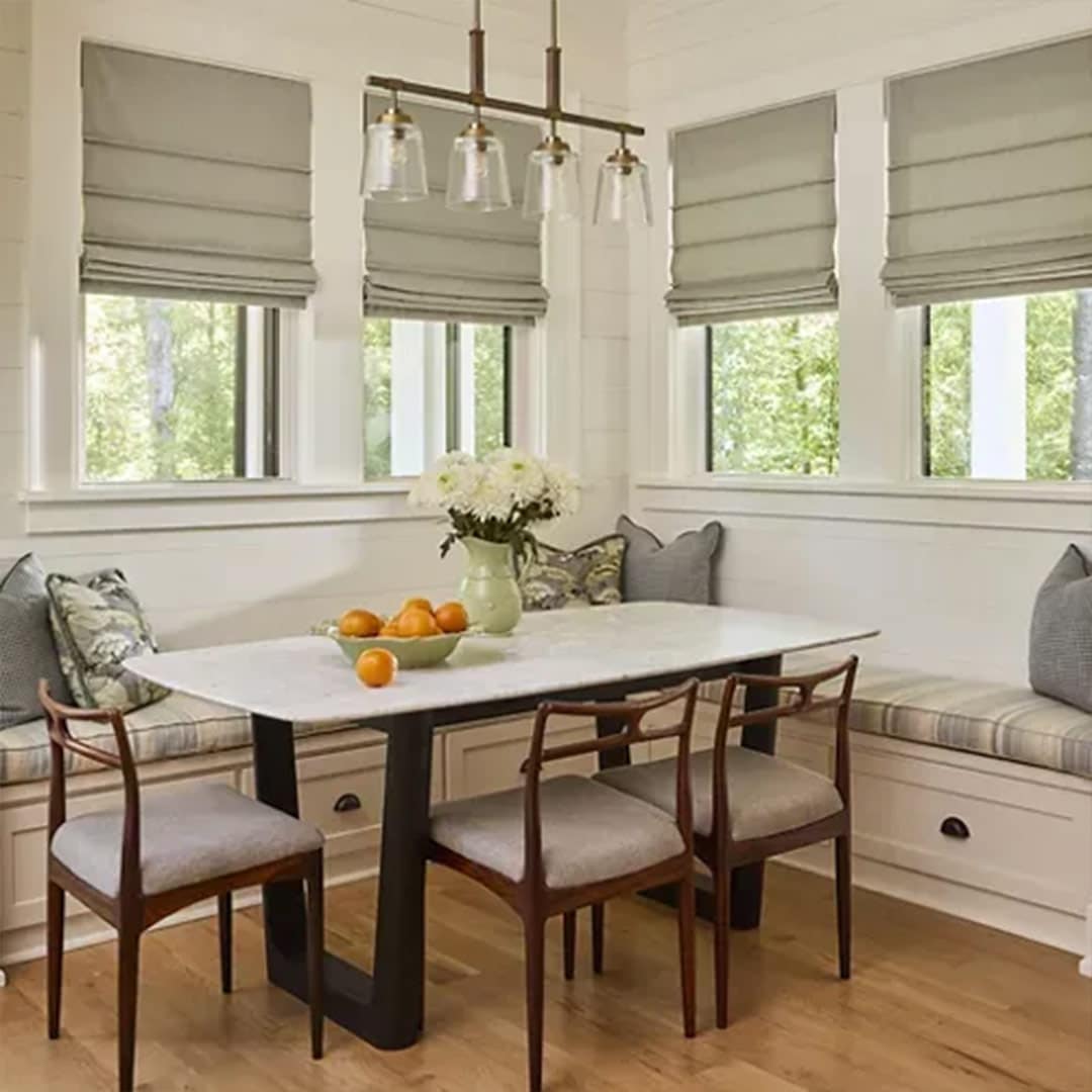 Cozy kitchen nook with built-in window seating, surrounded by large windows and Roman blinds, featuring a white dining table, wooden chairs, pendant lighting, and fresh flowers in a charming, light-filled space.