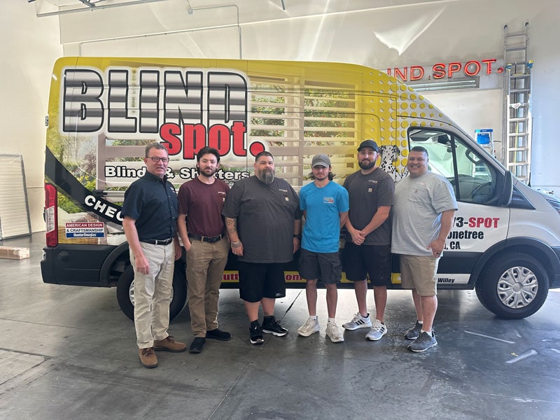 Group of five men standing in front of a Blind Spot vehicle inside a garage, smiling at the camera.