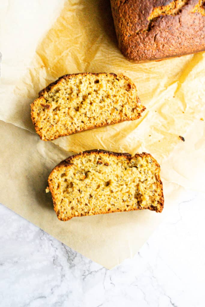 Two slices of orange poppyseed loaf on a piece of baking paper and a loaf in the background.