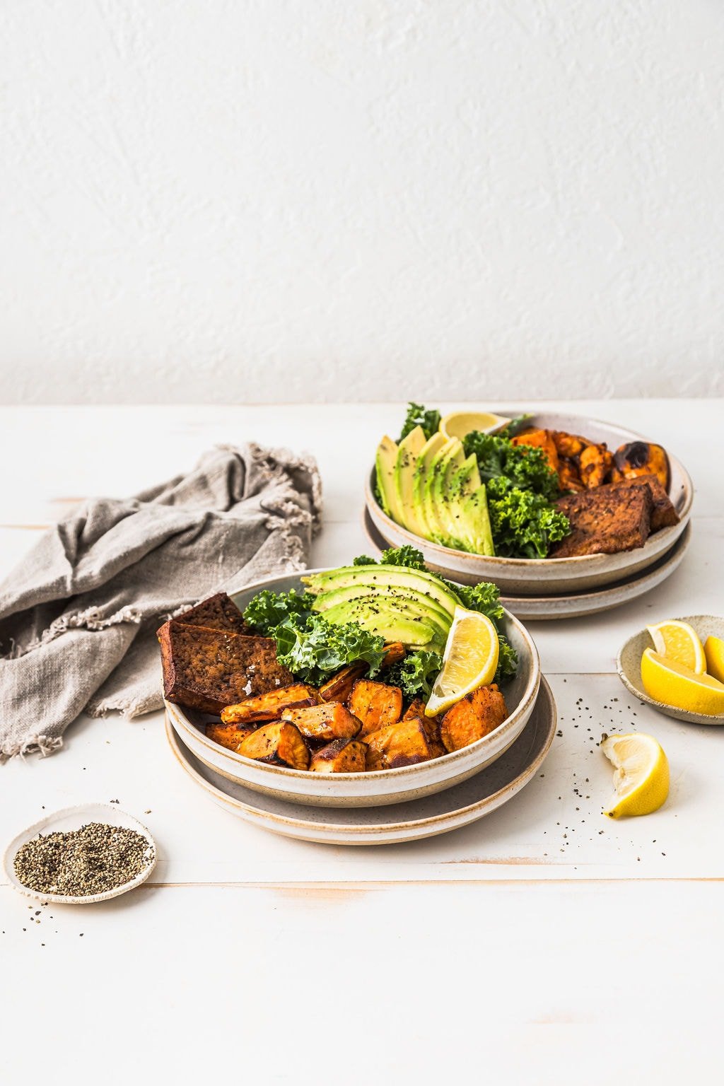 Front view of two sweet potato tofu nourish bowls on a white wooden table with petter, lemon and a grey napkin.