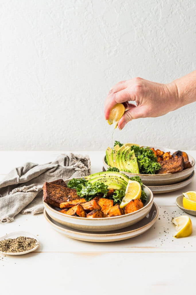Front view of a hand squeezing lemon juice over the top of a sweet potato tofu nourish bowl.