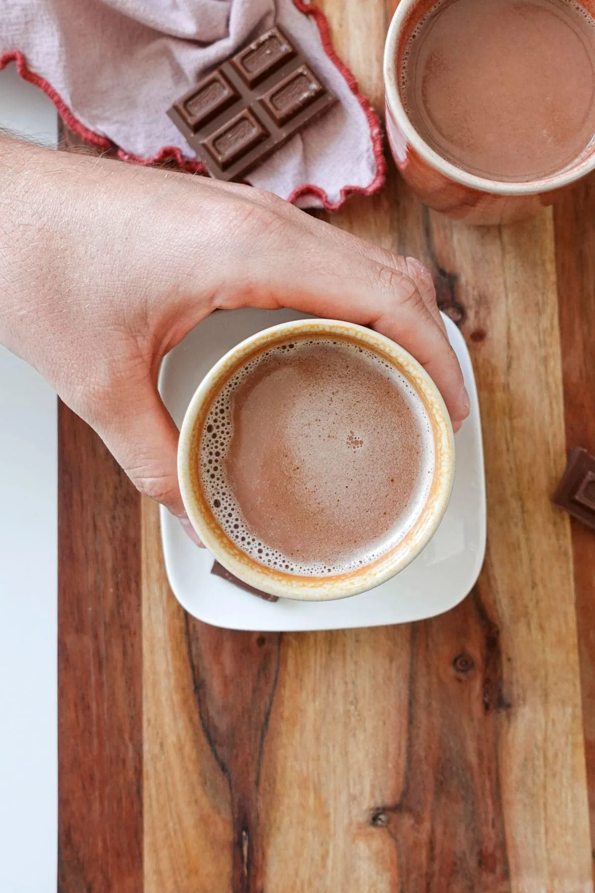 An overhead shot of a hand reaching in and grabbing a mug of Biscoff hot chocolate with another mug and chocolate pieces in the background.