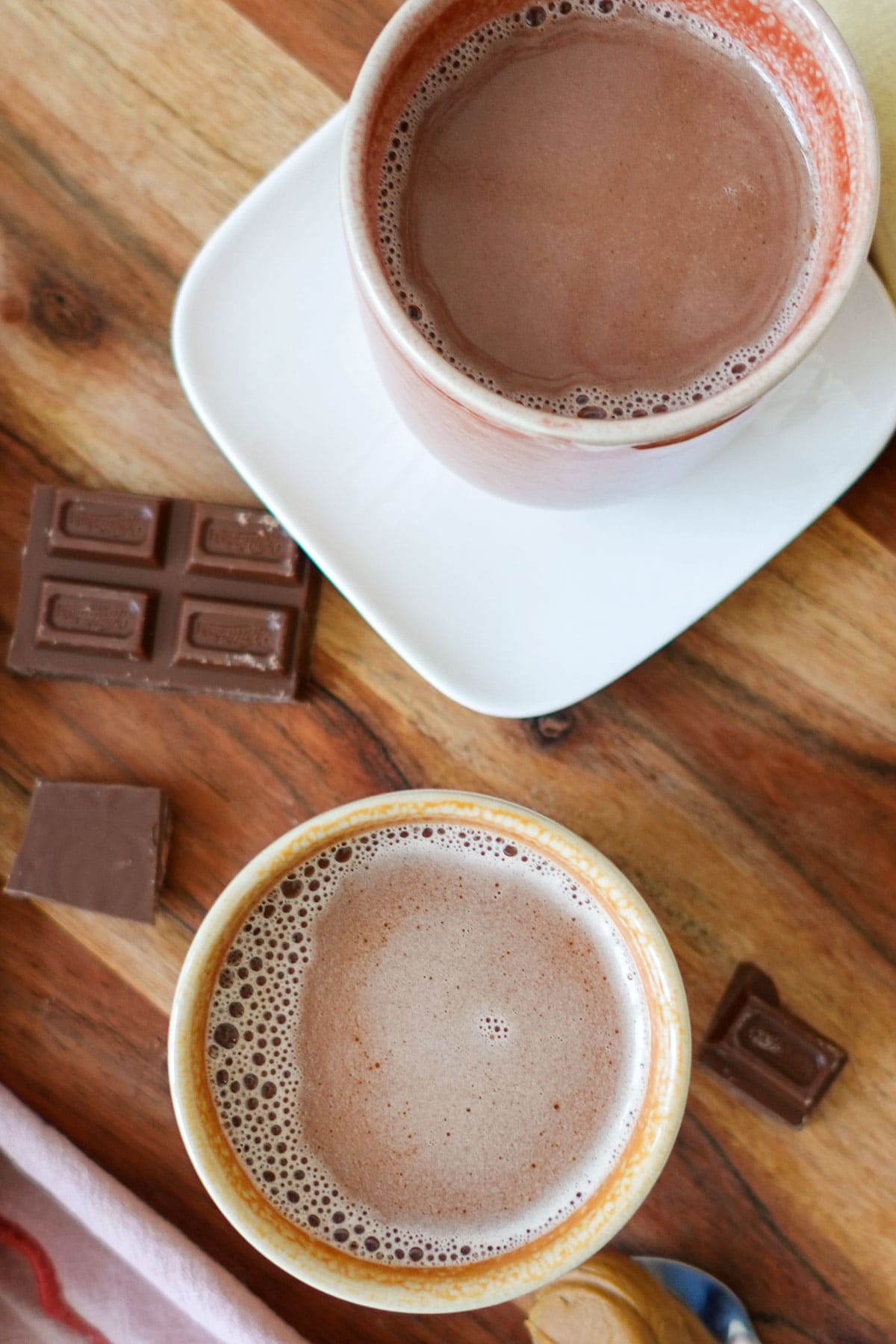 Overhead shot of two mugs of Biscoff hot chocolate on a wooden board with chocolate pieces all around.