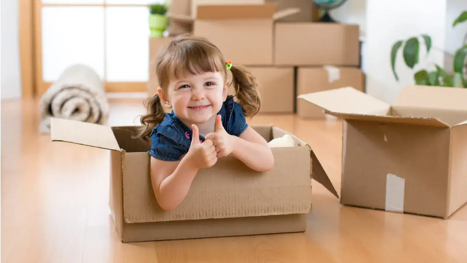 Smiling child giving thumbs up while sitting in a cardboard box amidst moving boxes, representing the joy of moving with kids.