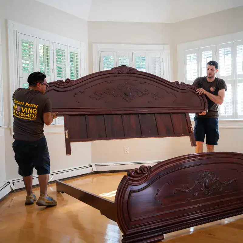 Two movers from Smart Penny Moving carefully lifting a wooden bed headboard inside a well-lit room, with a bed frame visible in the foreground.