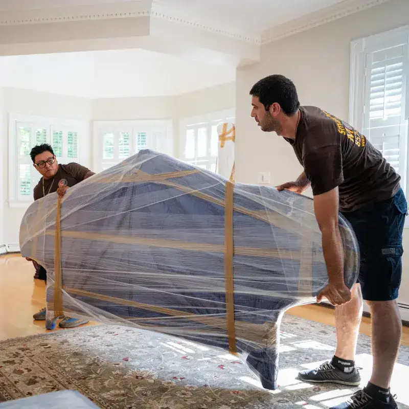 Two movers carefully lifting a wrapped sofa, demonstrating protective moving techniques in a well-lit living room.