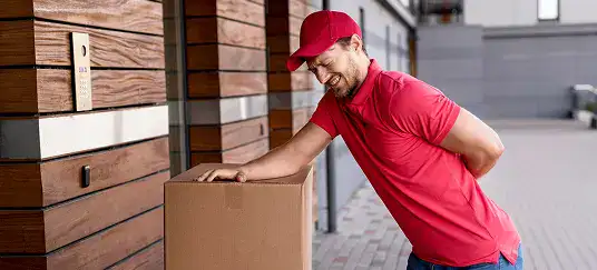 Mover in red shirt and cap, smiling while handling a large cardboard box, representing professional packing services for commercial moves.
