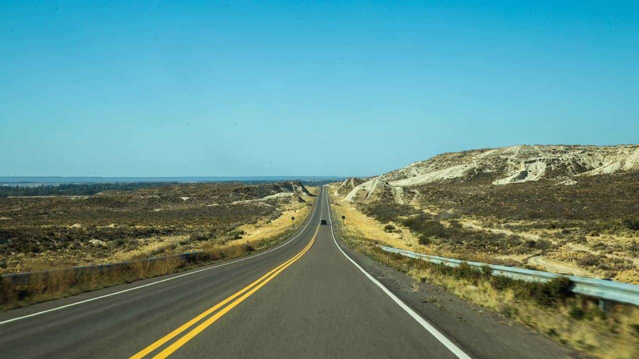 A straight two-lane highway stretches into the distance through a dry, hilly Patagonian landscape under a bright blue sky, with rugged terrain on both sides and no cars in sight.