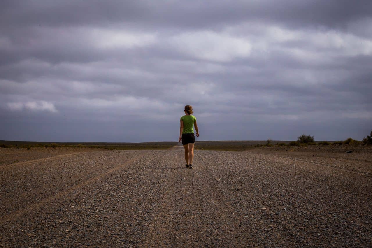 A woman in a green shirt and black shorts walks alone down a vast gravel road in the windswept plains of Patagonia. Heavy clouds loom above, adding to the sense of isolation and wild freedom.