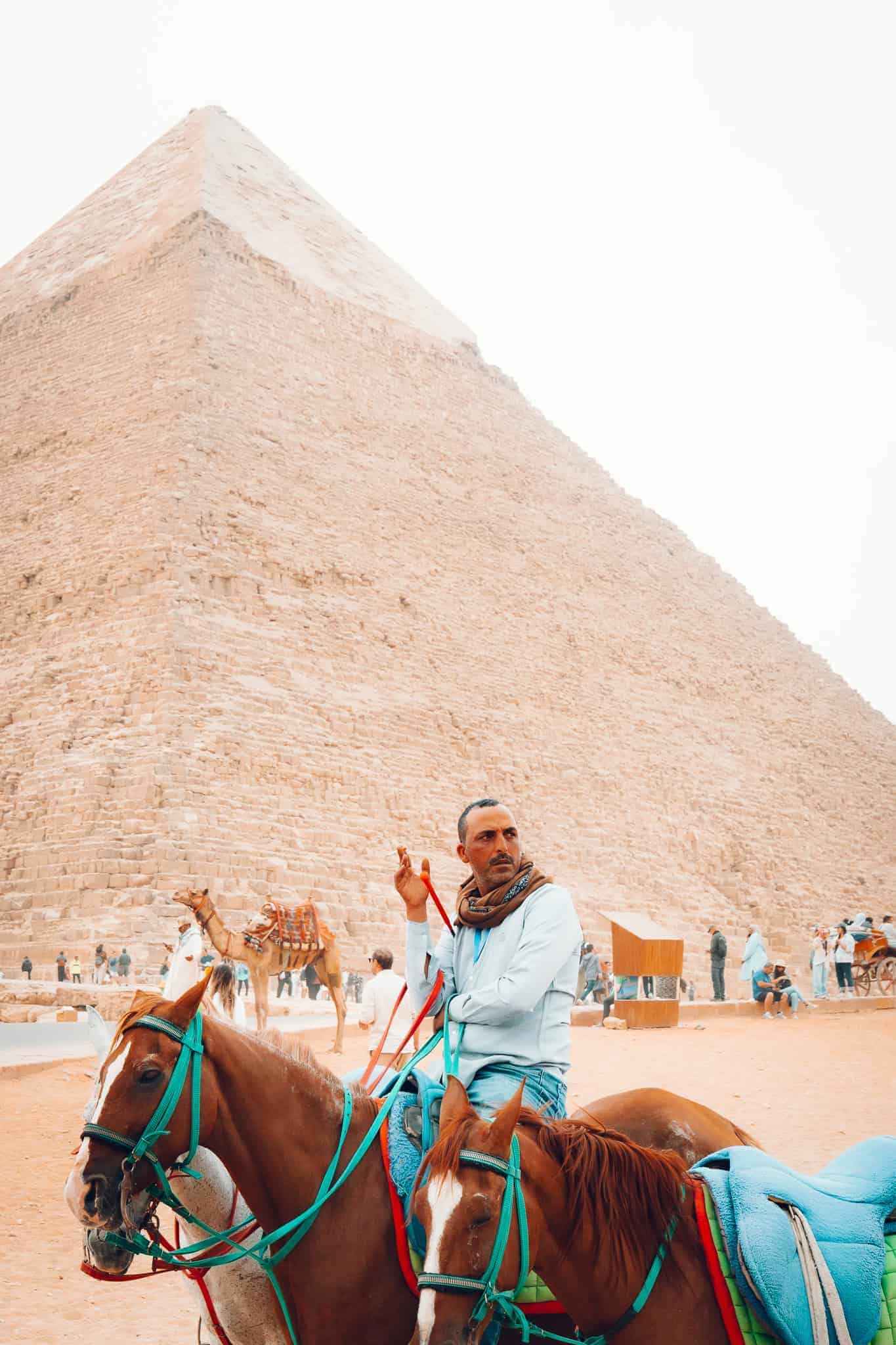 A man dressed in traditional attire sits on horseback in front of the towering Great Pyramid of Giza, surrounded by other riders and camels.
