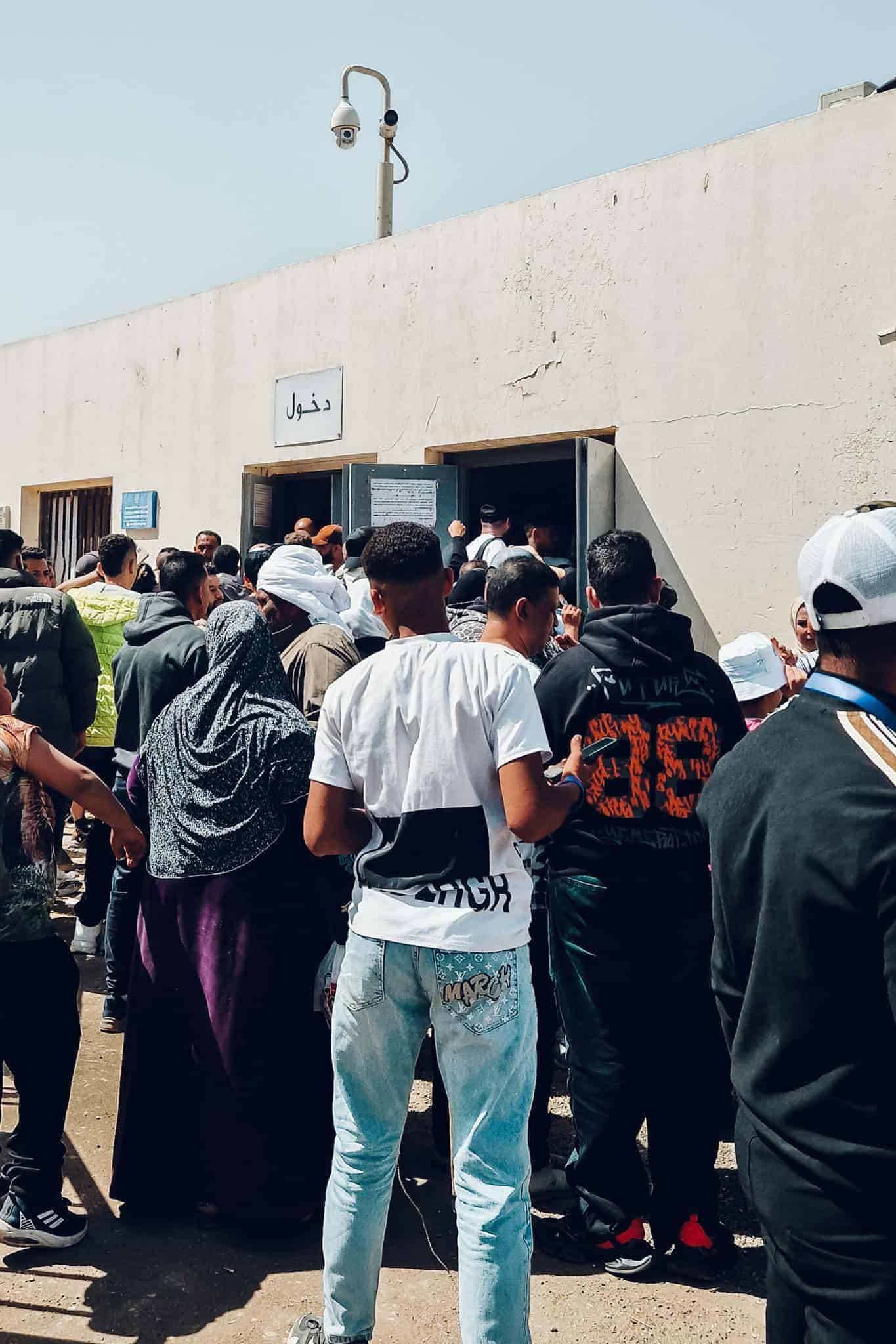 A crowd gathers outside a small white building marked with Arabic signs at a local access point to the pyramids.