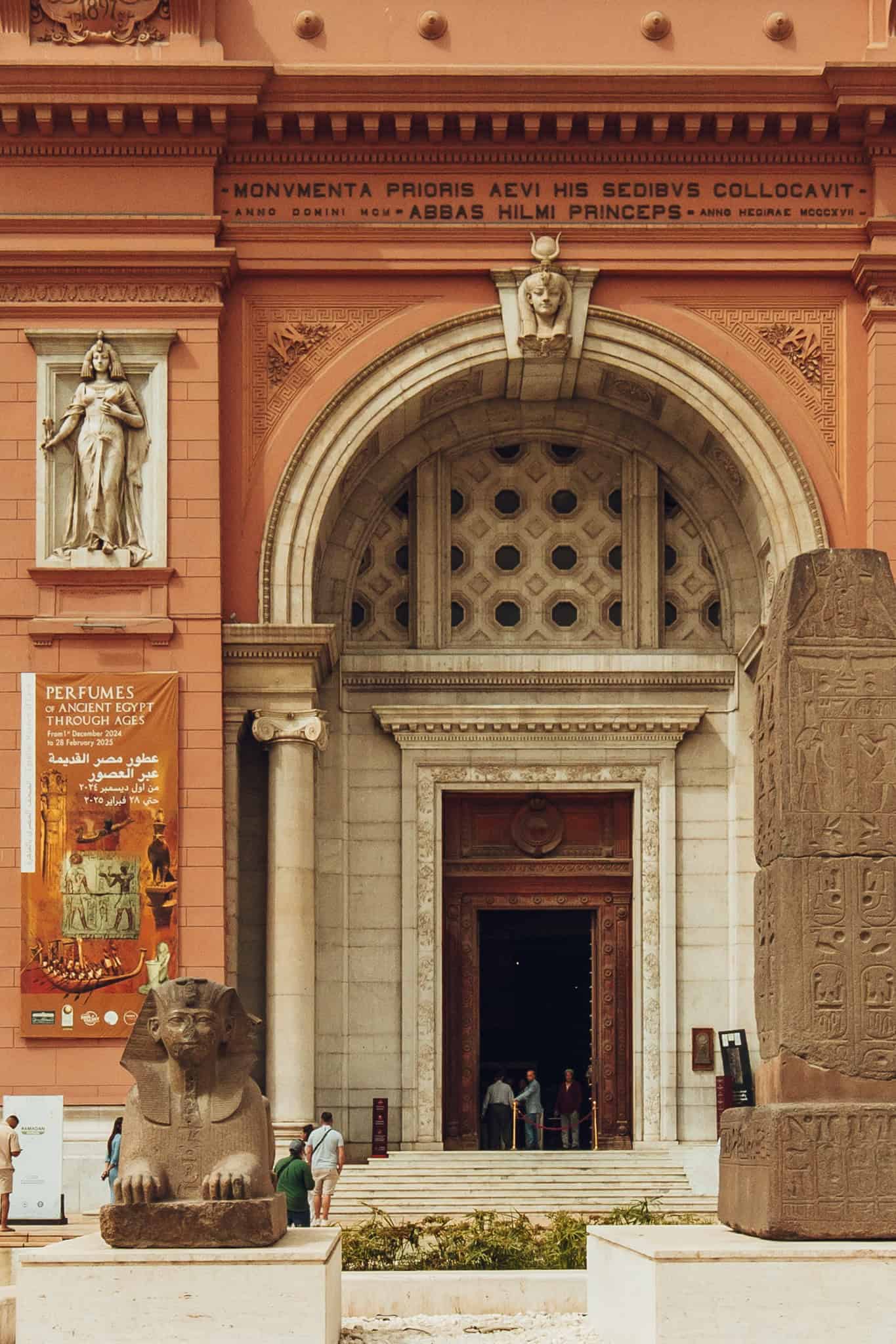 Front view of the entrance to the Old Egyptian Museum in Cairo, showcasing its grand arched doorway, neoclassical architectural details, and a seated sphinx statue in the foreground. A banner for the “Perfumes of Ancient Egypt” exhibition hangs beside the door, and a few visitors are seen entering the building.