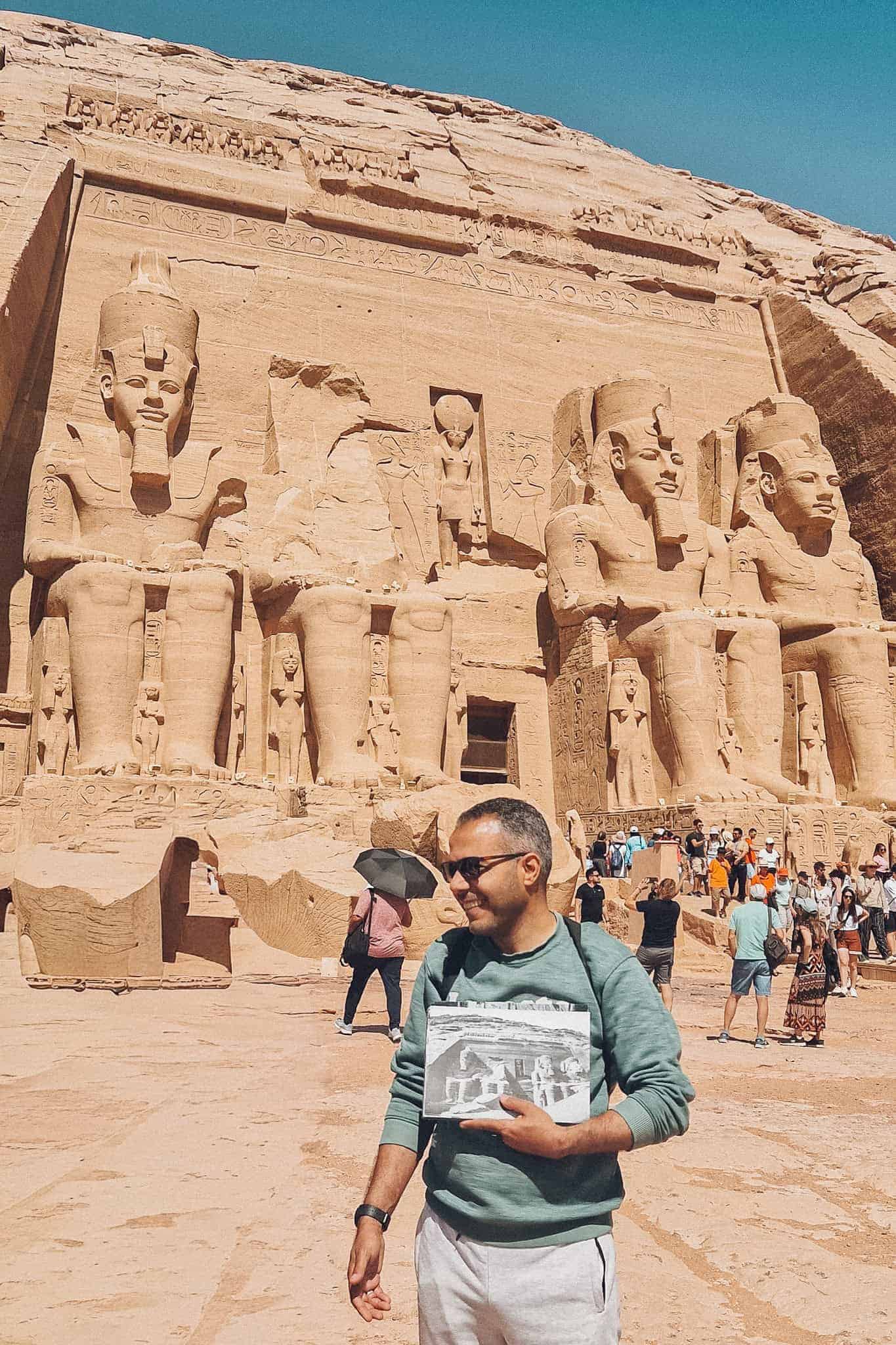 A smiling man stands in front of the colossal statues at the entrance of Abu Simbel’s Great Temple, holding a historical black-and-white photo showing the original temple site before relocation.