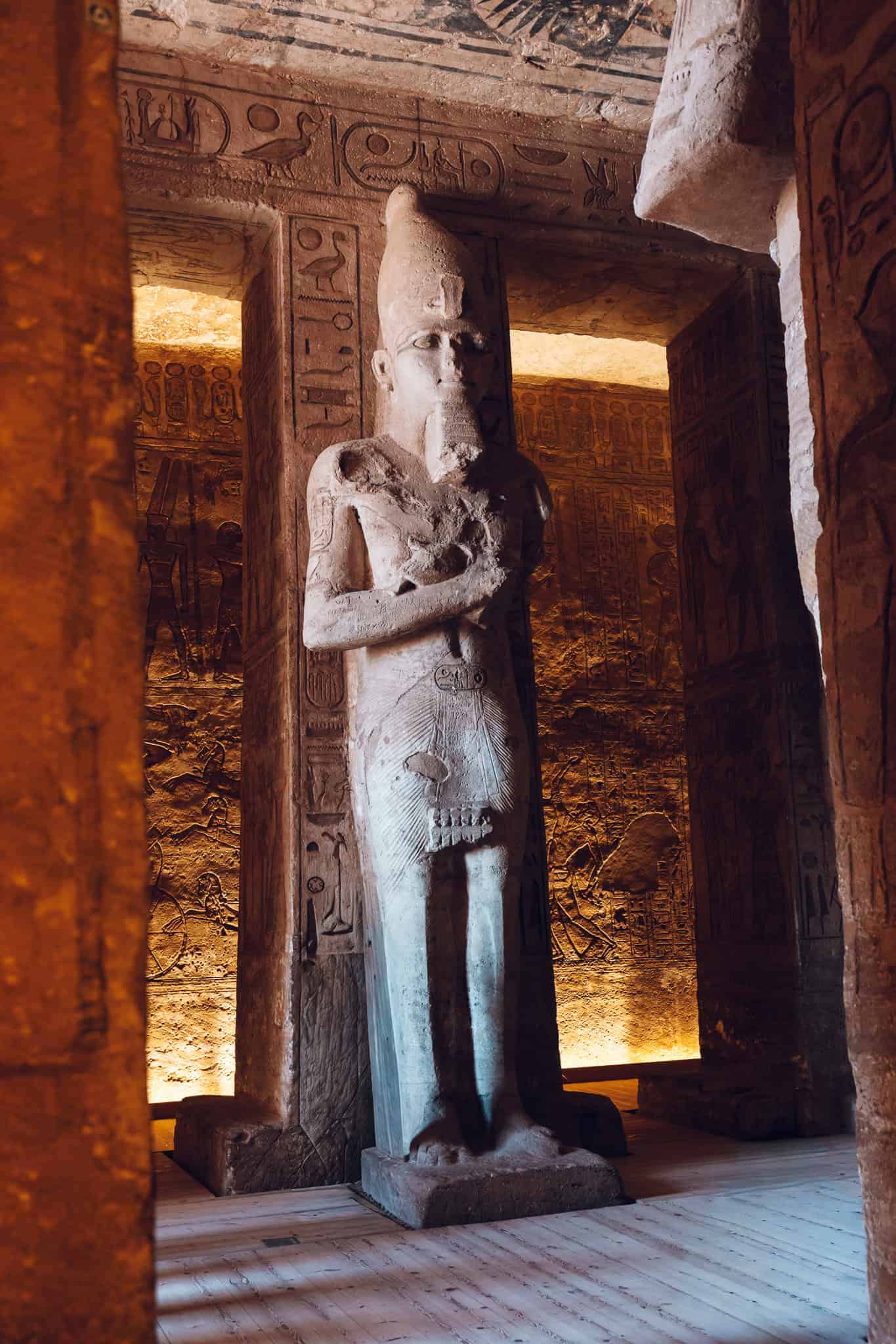 Full-body statue of Ramesses II standing against a hieroglyph-covered pillar inside Abu Simbel, softly lit by warm interior lighting.
