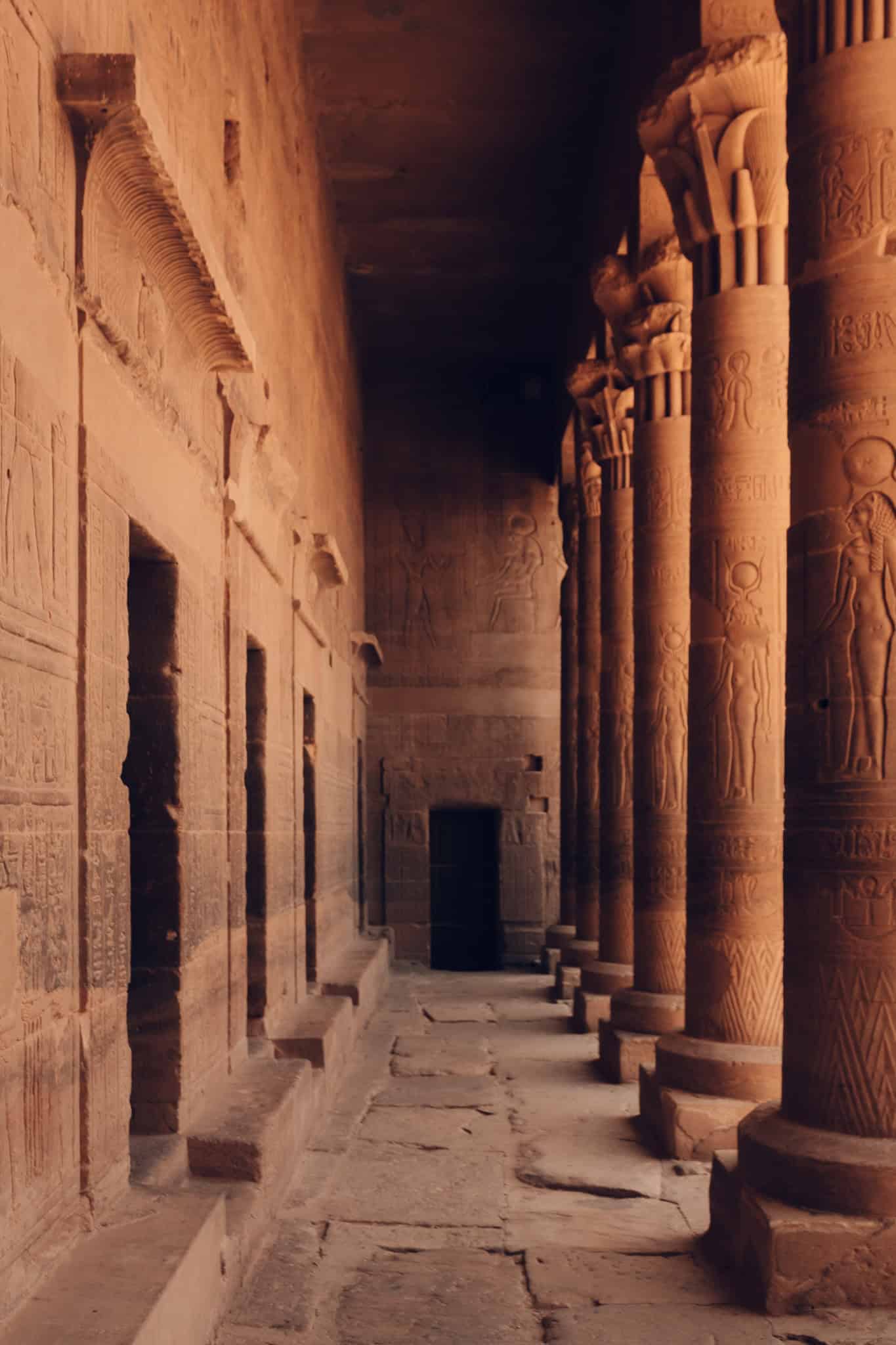 Interior hallway at Philae Temple in Aswan, Egypt, with a long row of carved sandstone columns and faded wall reliefs lit by soft ambient light.