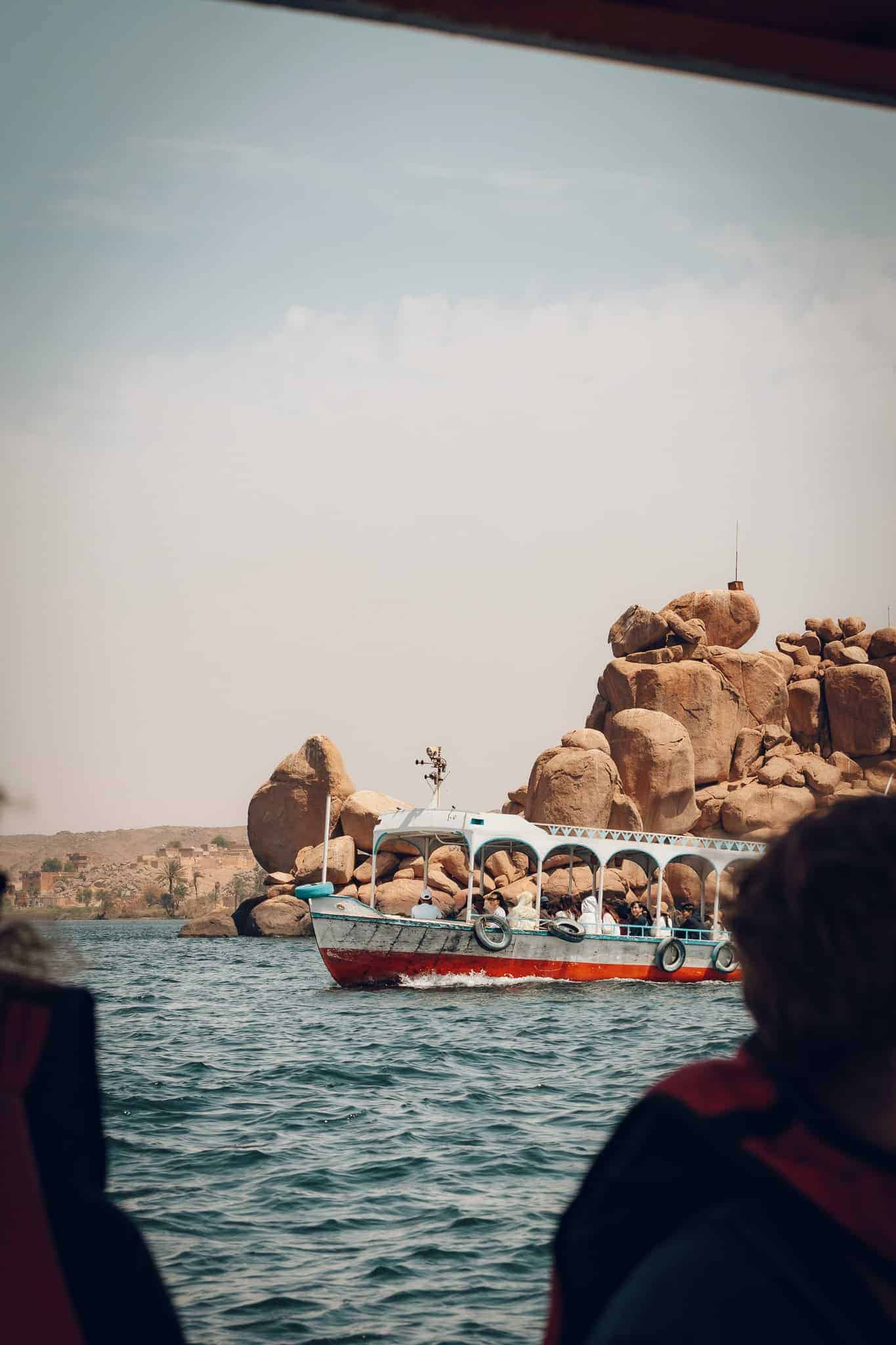 A small ferry boat cruises past massive boulders on the Nile River in Aswan, Egypt, carrying passengers under a hazy sky while framed by other travelers onboard.