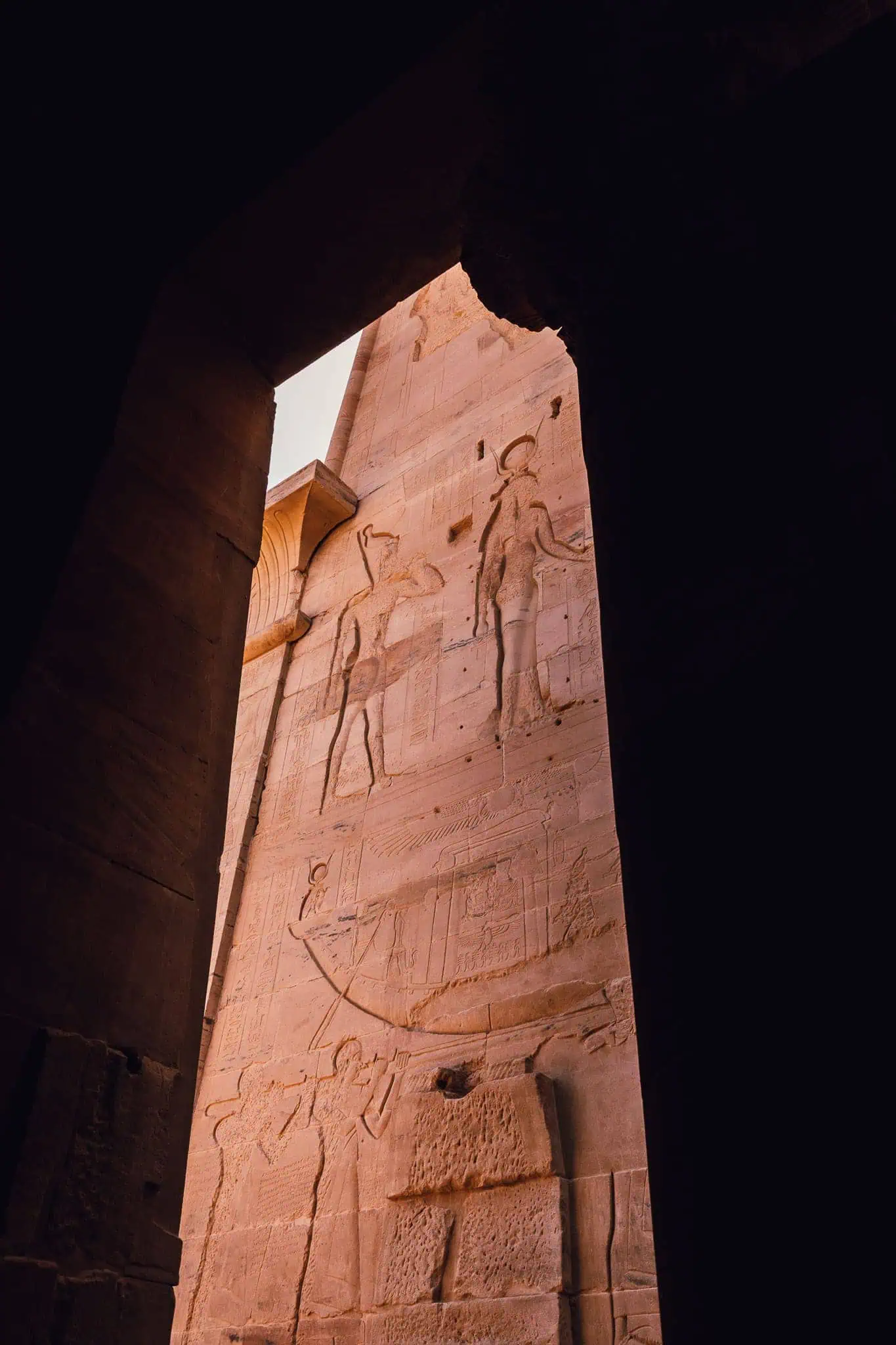 View through a shadowed temple entrance at Philae Temple in Aswan, Egypt, framing a tall pylon wall with large carvings of gods and pharaohs in sunlight.