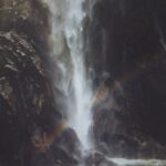 Waterfall in Milford Sound with rainbow and rainforest backdrop.
