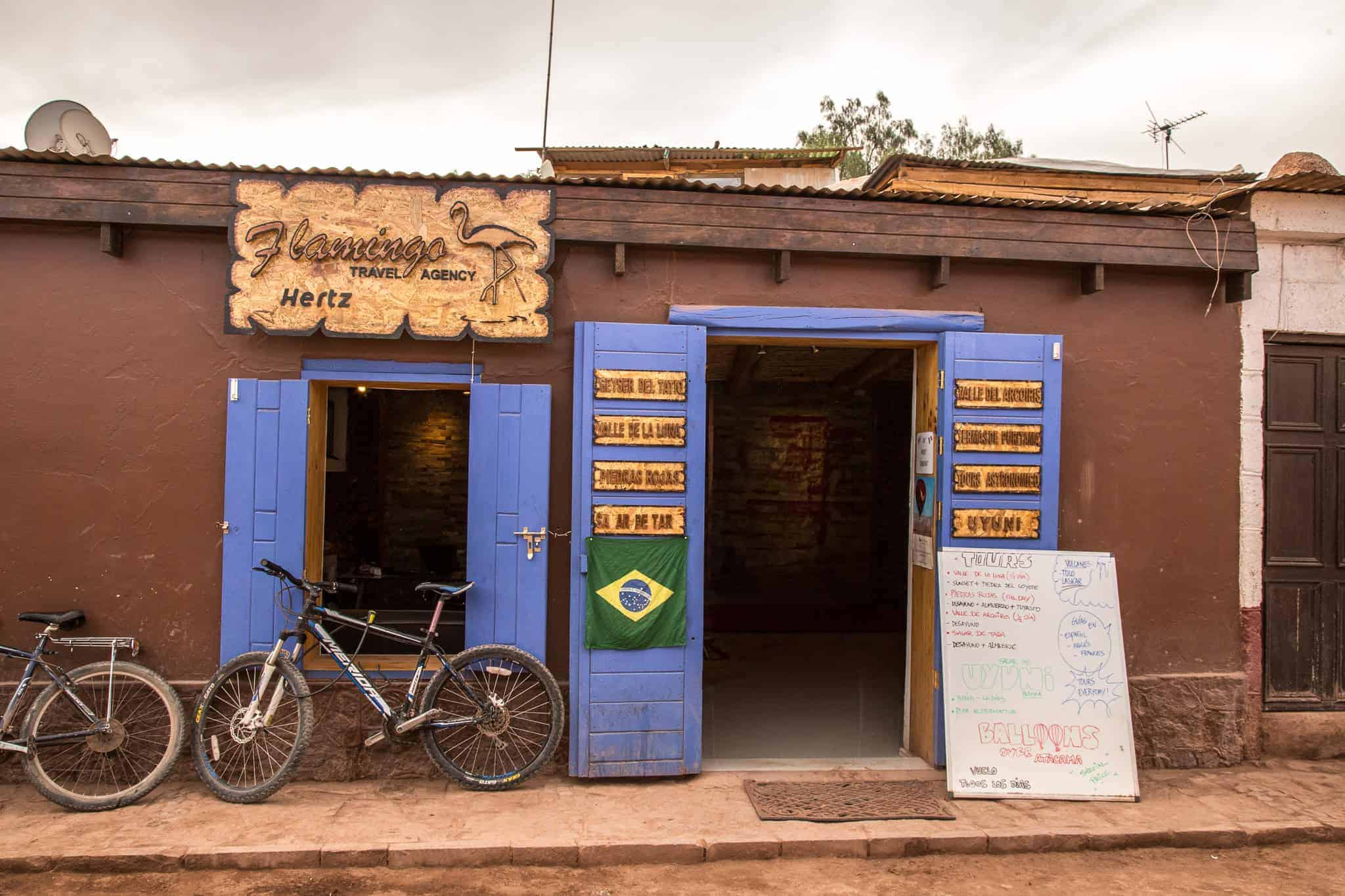 A rustic travel agency with bright blue doors and a wooden sign reading "Flamingo Travel Agency" adorned with a flamingo illustration, flanked by parked bicycles and a Brazilian flag hanging by the entrance.
