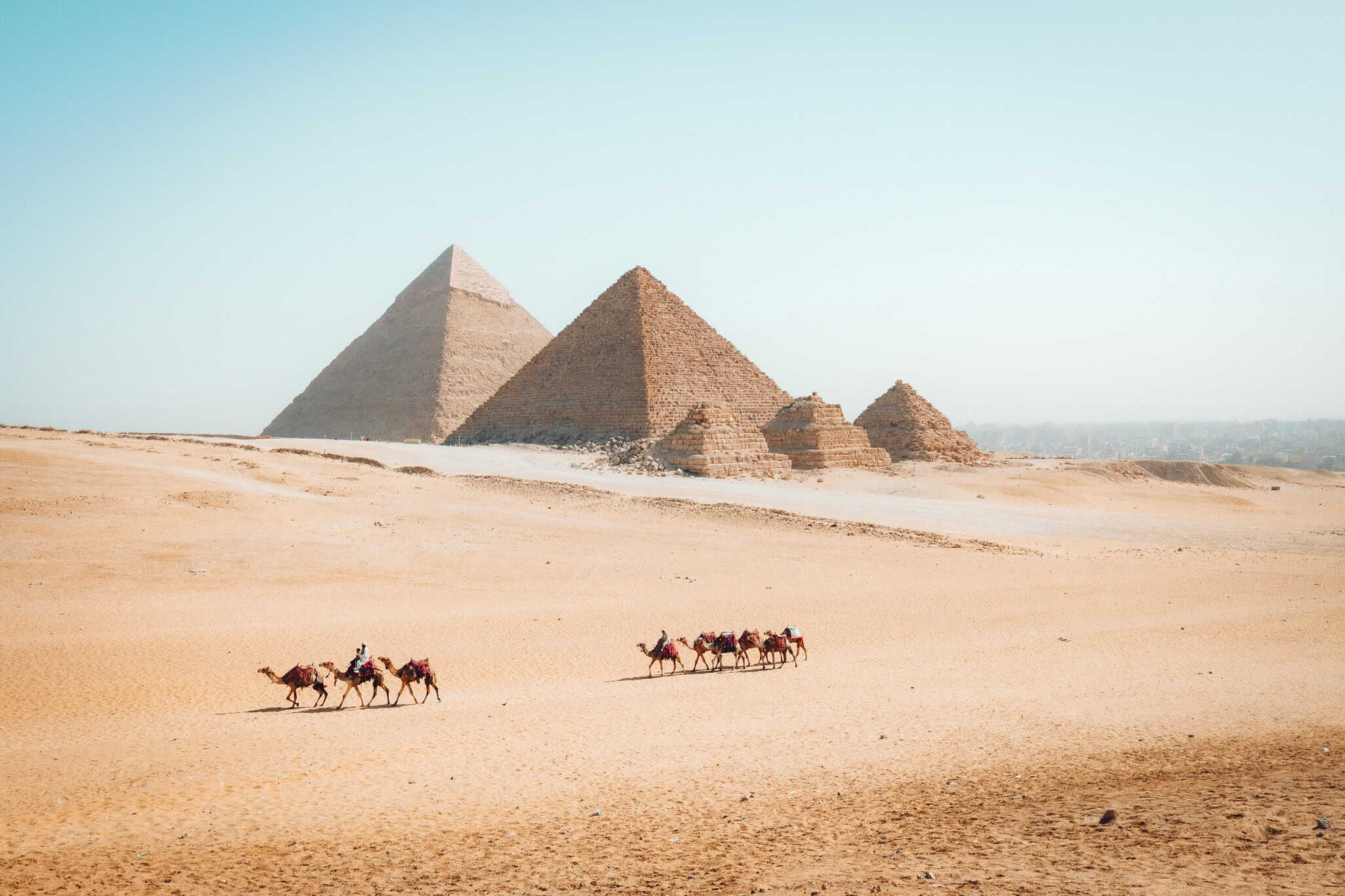 Camel riders traverse the desert in front of the pyramids of Giza, with sunlight casting strong shadows and highlighting the ancient stone structures.