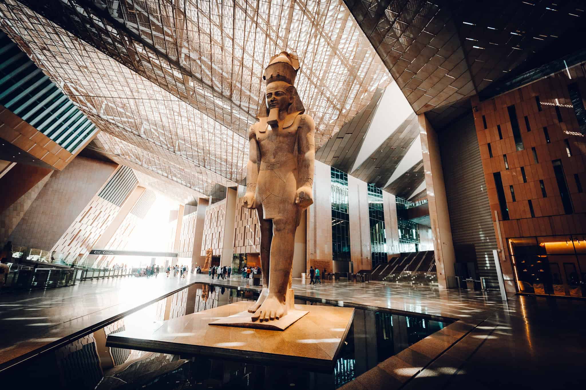 Massive statue of Ramses II stands proudly inside the sunlit atrium of the Grand Egyptian Museum. The modern architecture features soaring glass and metal angles with dramatic lighting.