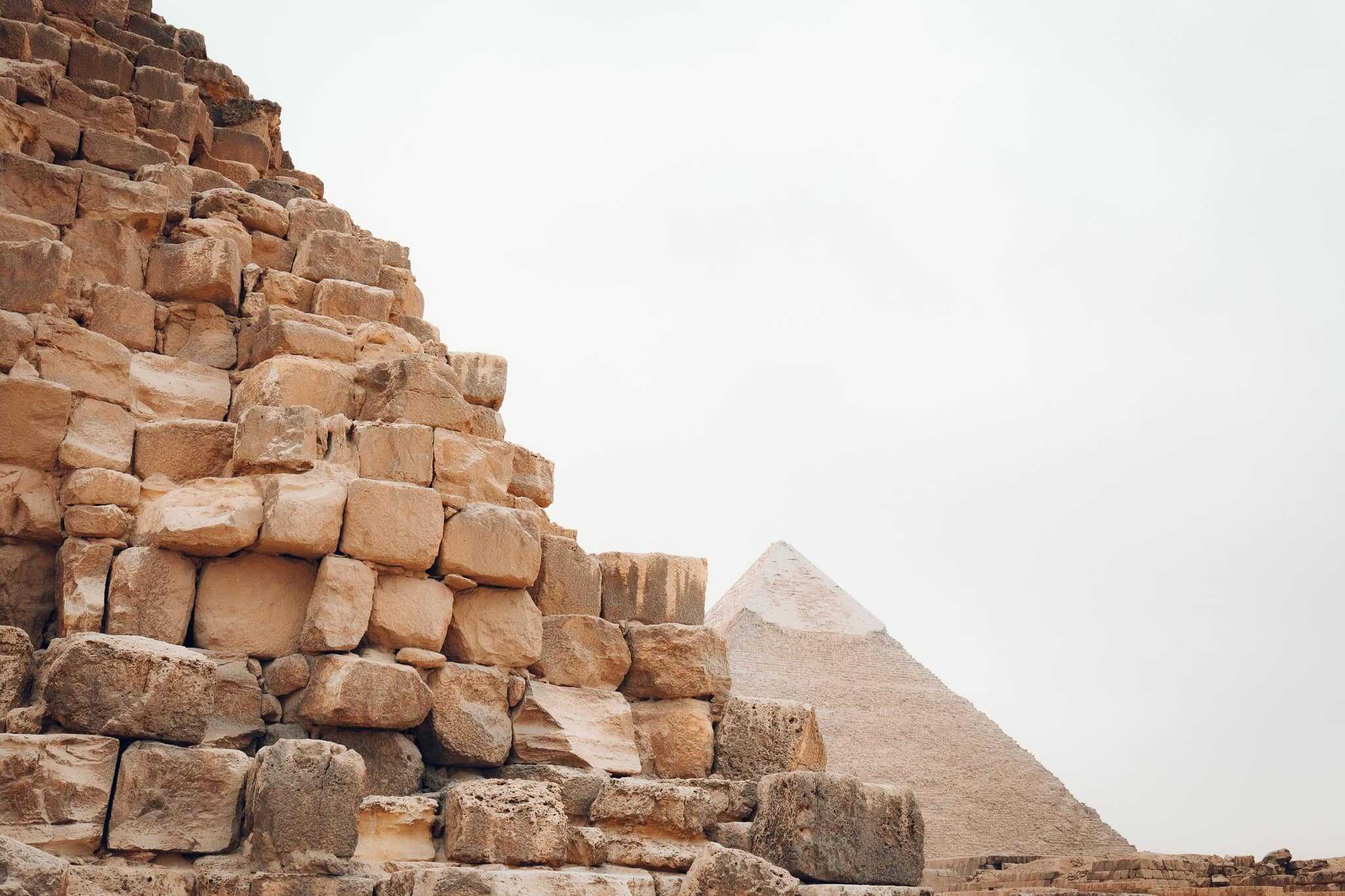 Close-up of rugged stone blocks from the Pyramid of Khufu in the foreground, with the Pyramid of Khafre peeking over in the distance under a pale sky.