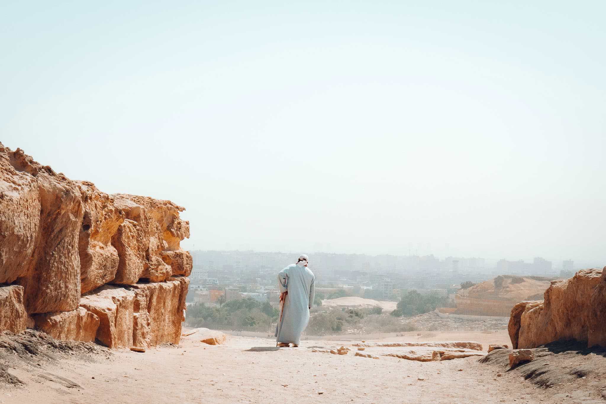 A man in traditional Egyptian dress walks through a natural rock corridor, overlooking Cairo's hazy skyline from the Giza Plateau.