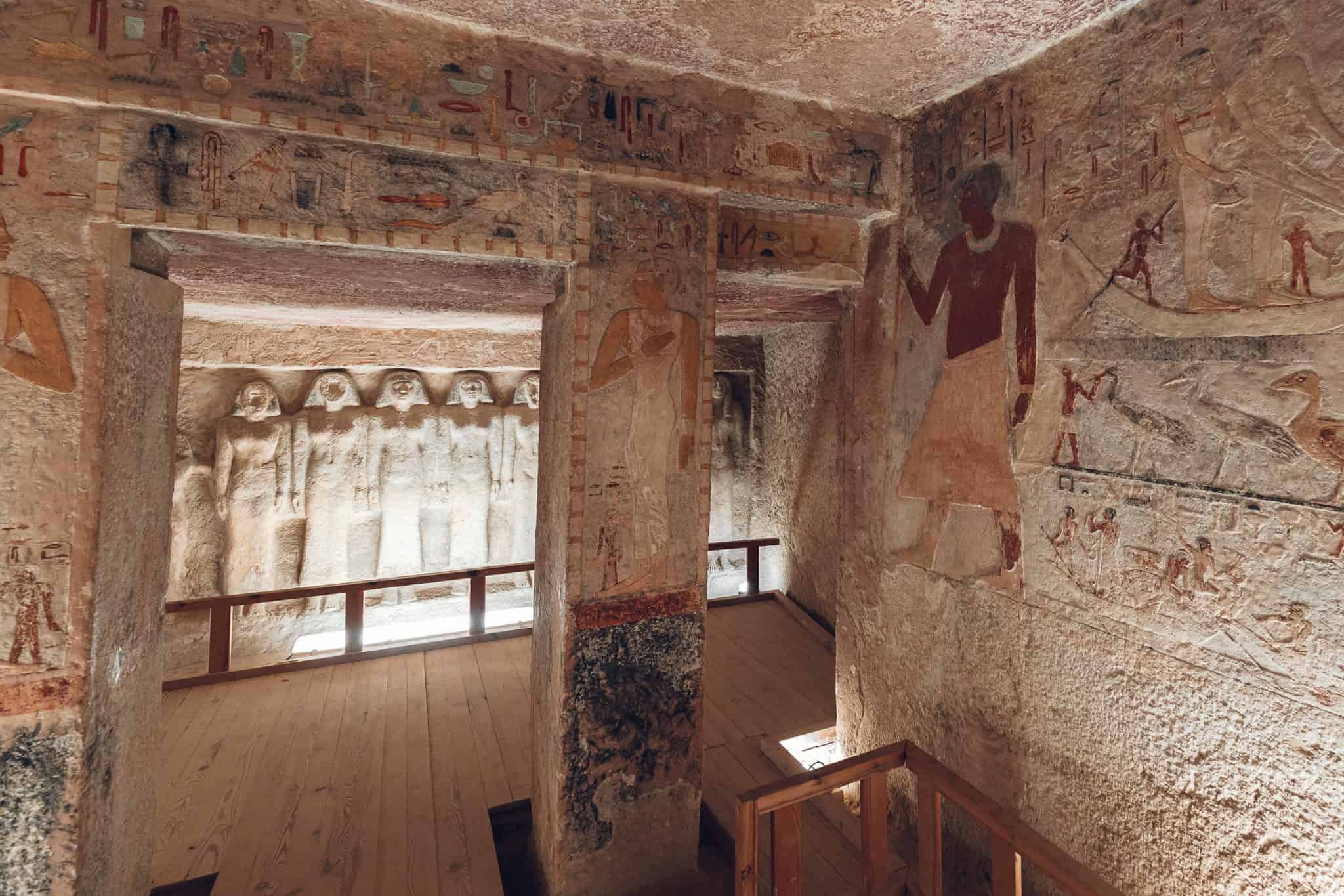 Inside the tomb of Queen Meresankh III, detailed wall carvings and faded yet vibrant paintings depict daily life and ceremonial scenes, with statues of figures lining the far wall. Soft lighting highlights the textures of the ancient stone and preserved pigments.