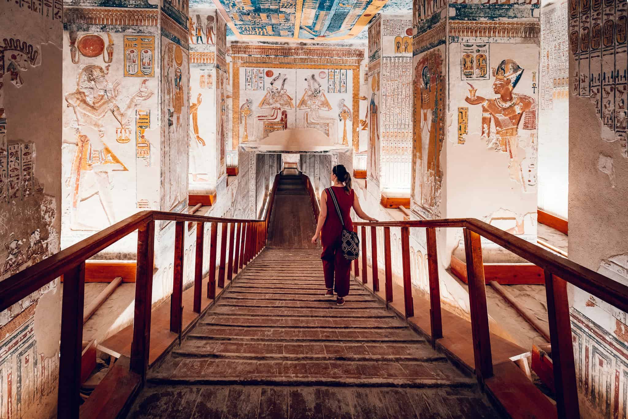 A woman walks down a wooden ramp into a vividly painted ancient tomb in the Valley of the Kings, surrounded by columns covered in Egyptian art and hieroglyphs.
