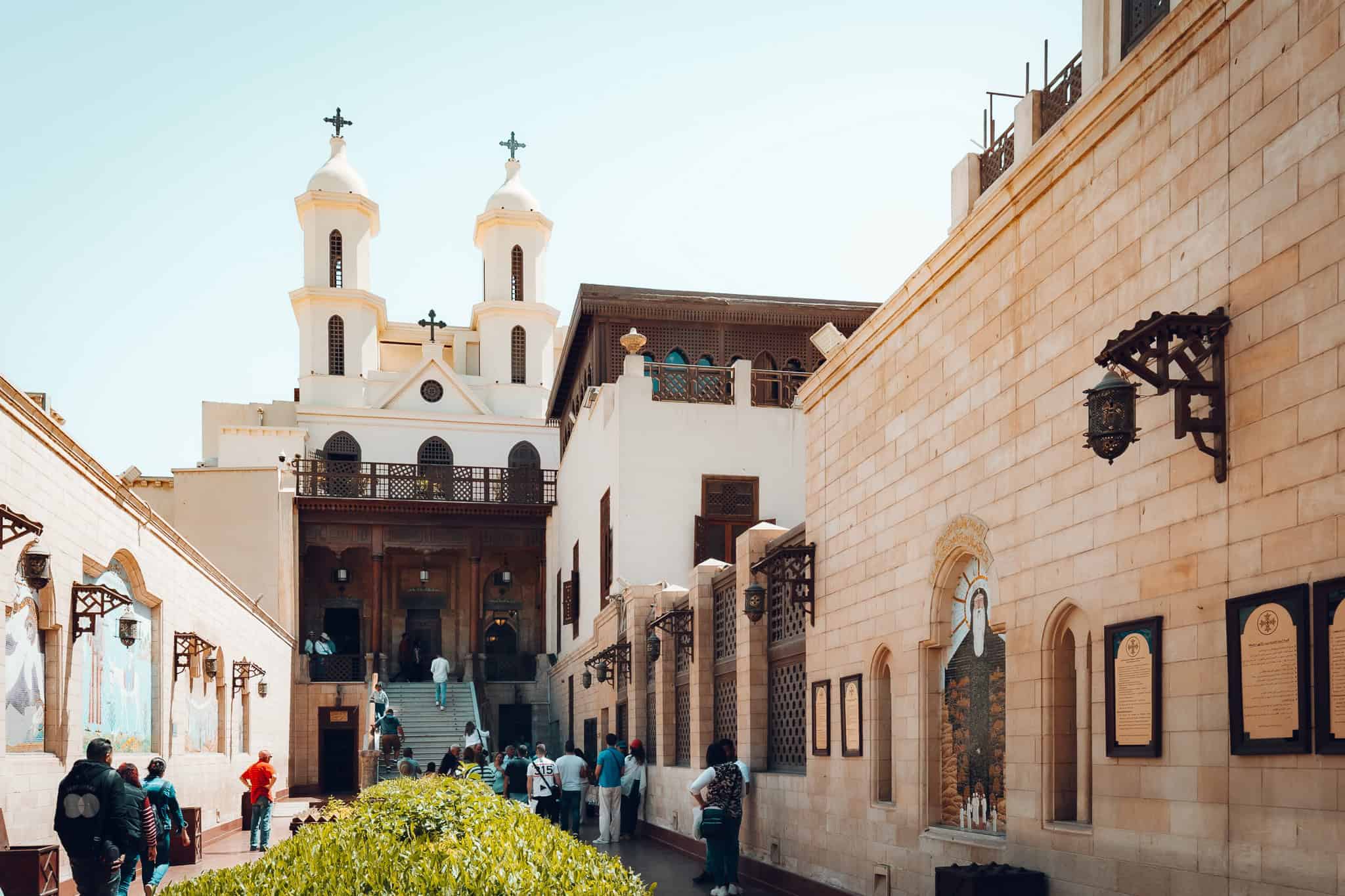 A view of the Hanging Church in Cairo, Egypt, showcasing its iconic twin bell towers and ancient Coptic architecture. Visitors are seen walking along the courtyard, surrounded by stone walls adorned with religious artwork and plaques.