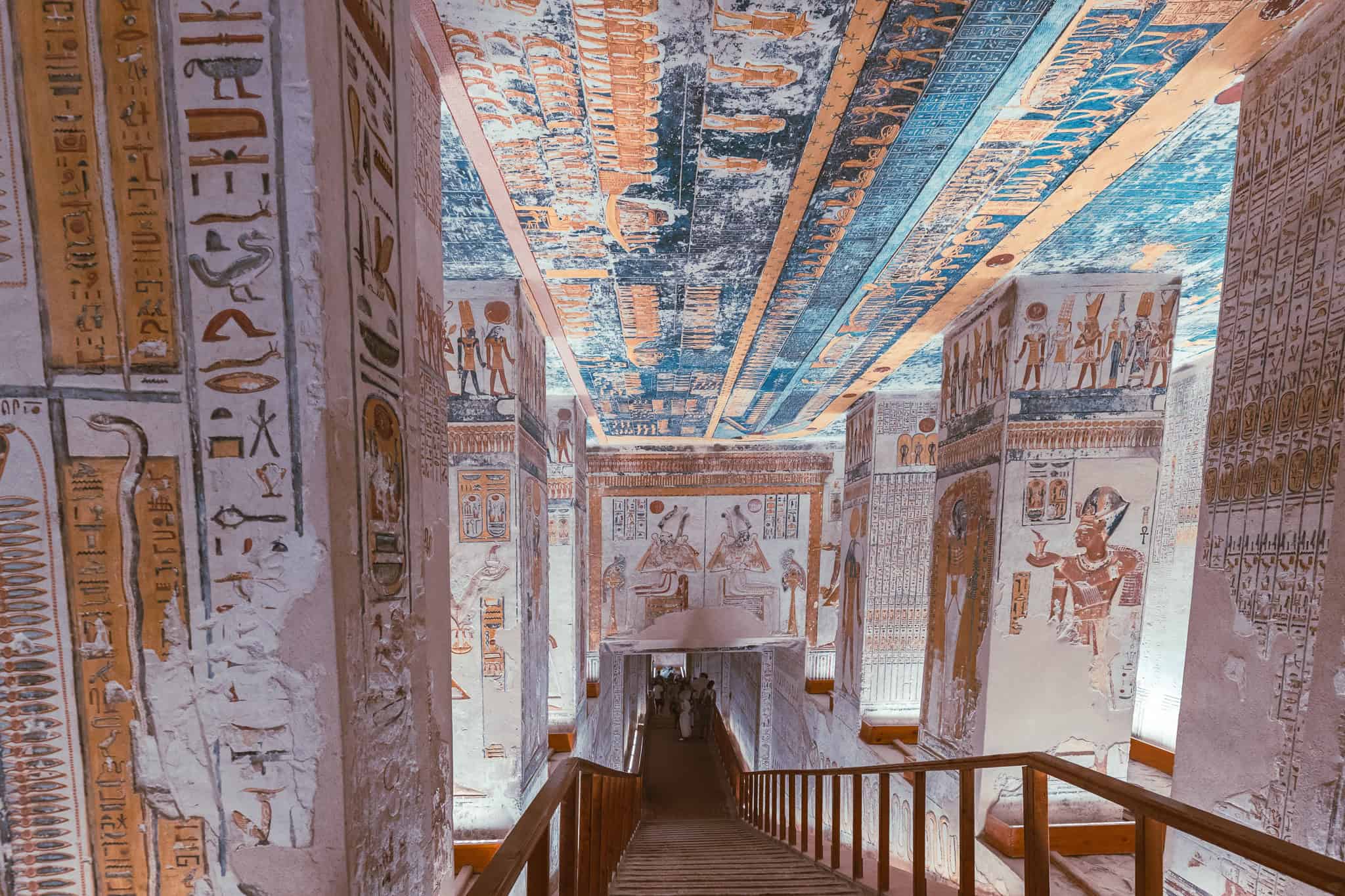 vividly decorated tomb corridor in the Valley of the Kings, Egypt, adorned with intricate hieroglyphs and colorful murals depicting scenes of Egyptian mythology. The ceiling features celestial artwork in vibrant blues and golds, while the walls display gods, pharaohs, and ceremonial rites. A wooden staircase leads visitors deeper into the sacred tomb, reflecting the grandeur of ancient Egyptian burial practices.