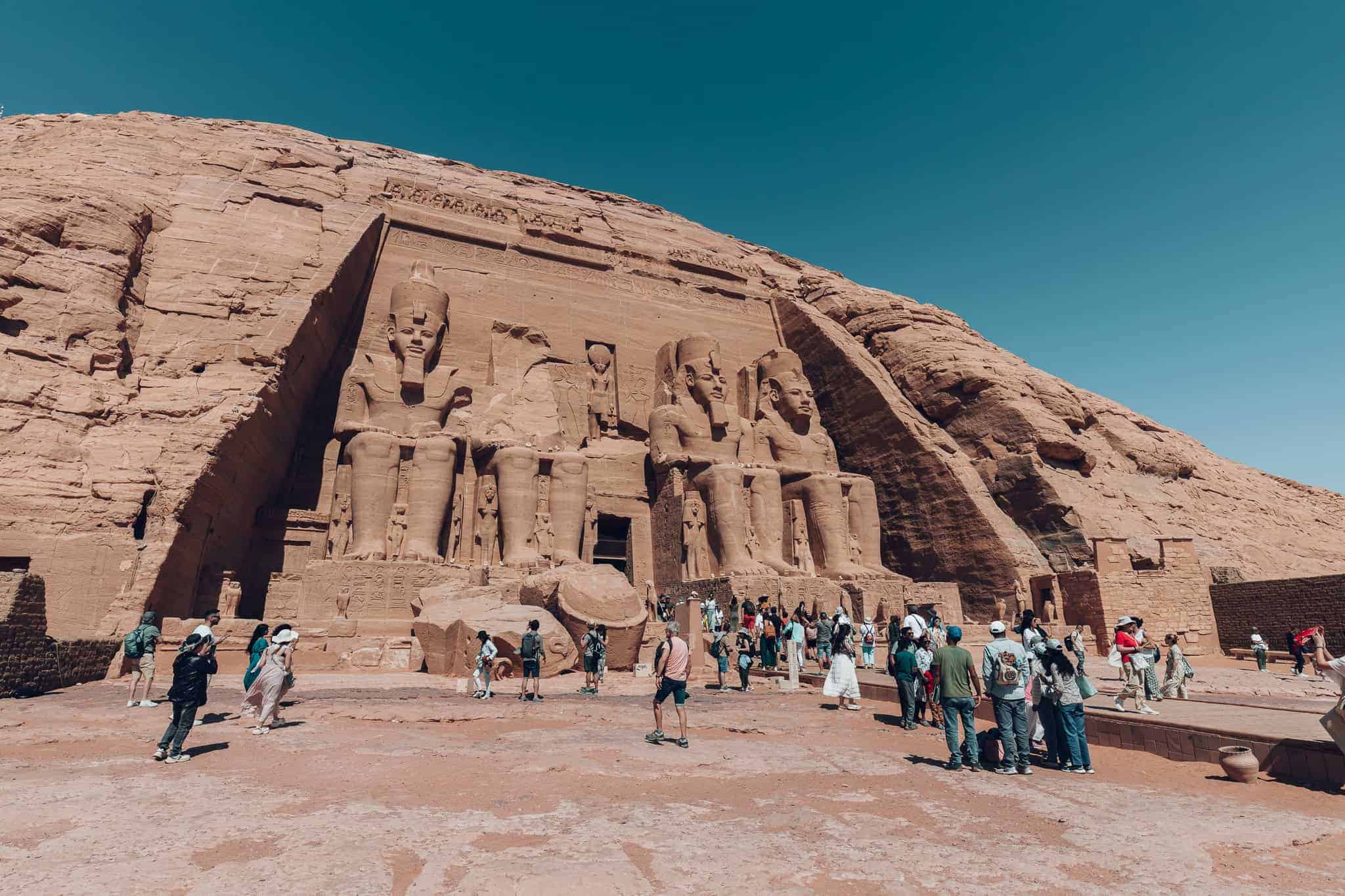 Tourists explore the forecourt of the Great Temple at Abu Simbel, standing in front of the enormous statues carved into the cliff under the bright desert sun.