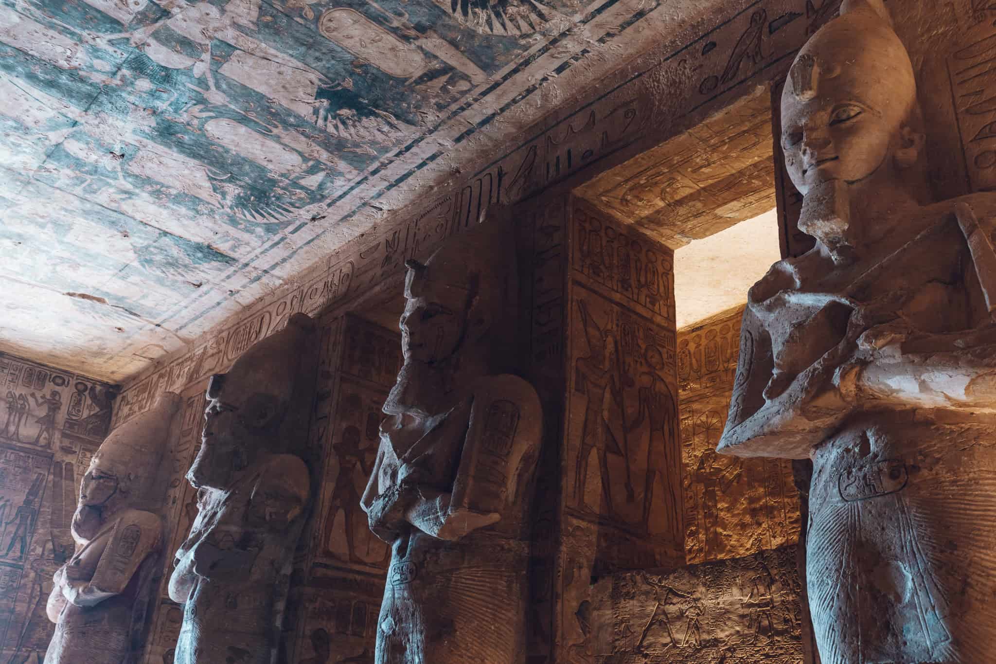 Interior row of monumental statues of Ramesses II in Abu Simbel, illuminated by warm lighting and framed by painted ceilings and carved pillars.