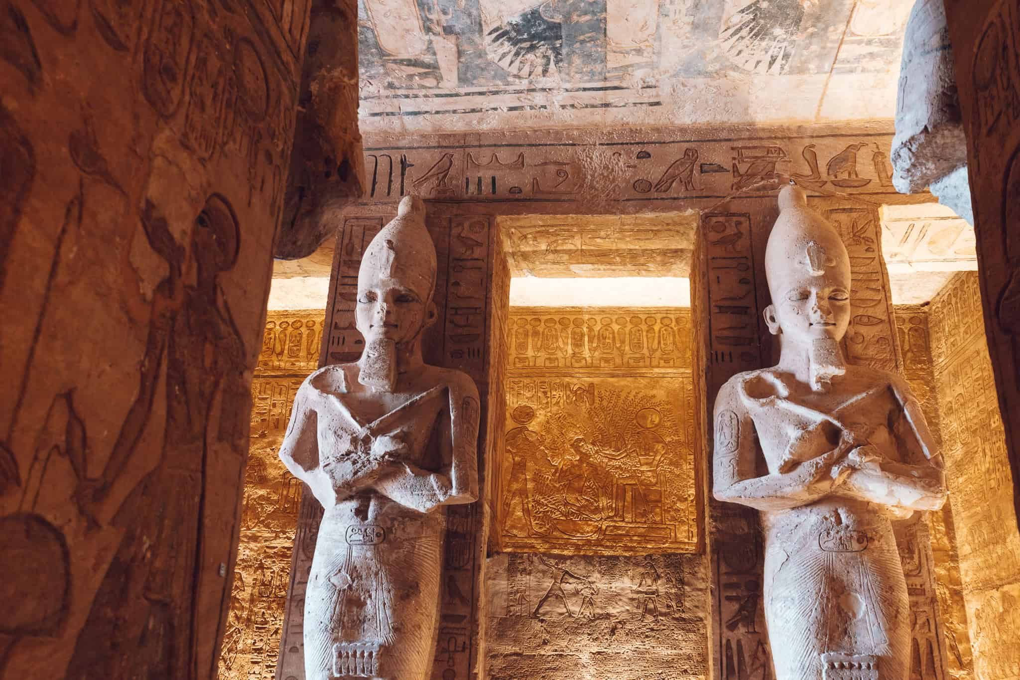 Two statues of Ramesses II with crossed arms flank an interior passage of Abu Simbel, with illuminated wall carvings and ceiling artwork in the background.