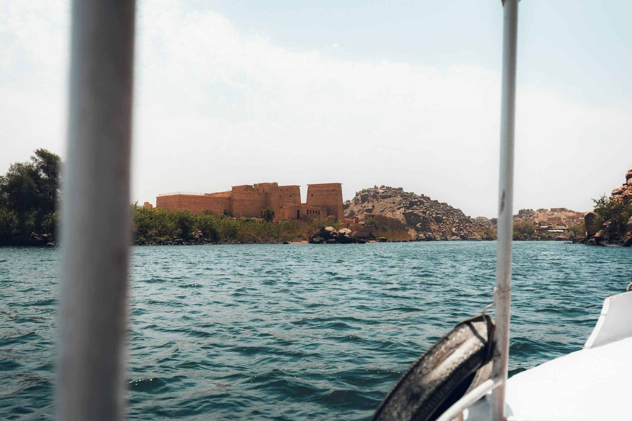 A boat ride approaches Philae Temple in Aswan, Egypt, with temple pylons and rocky hills in the distance, framed by a white metal boat railing in the foreground.