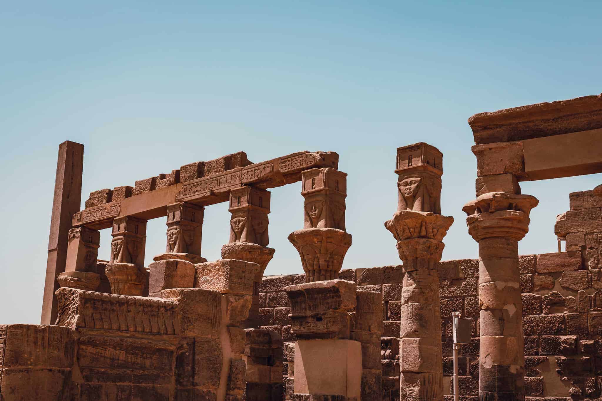 Close-up of Hathor-headed columns at Philae Temple in Aswan, Egypt, with carved capitals and stone lintels bearing hieroglyphs under a clear sky.