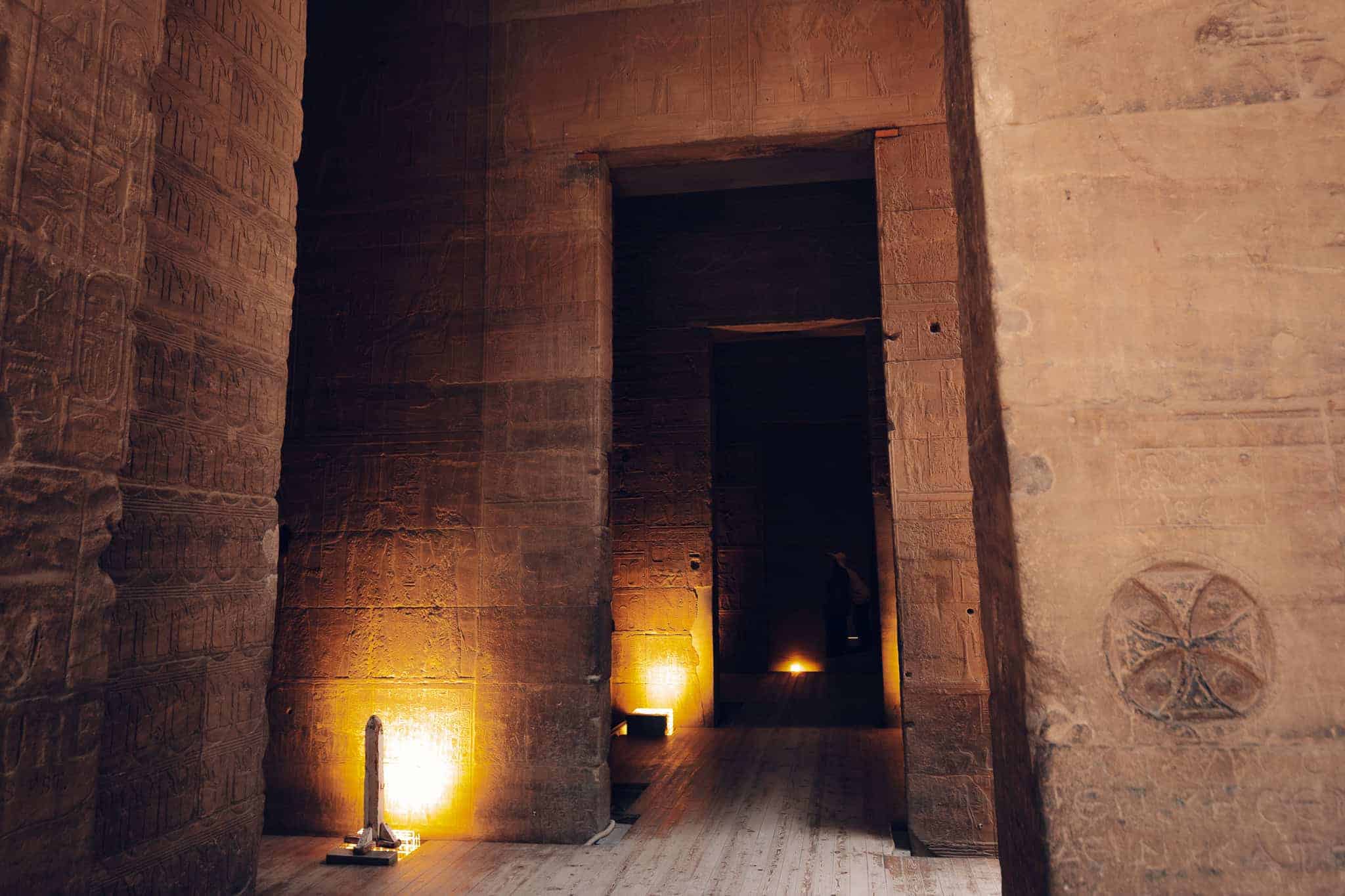 Soft lighting highlights hieroglyph-covered walls and a small lit obelisk replica inside Philae Temple in Aswan, Egypt, within a series of dark sandstone chambers.