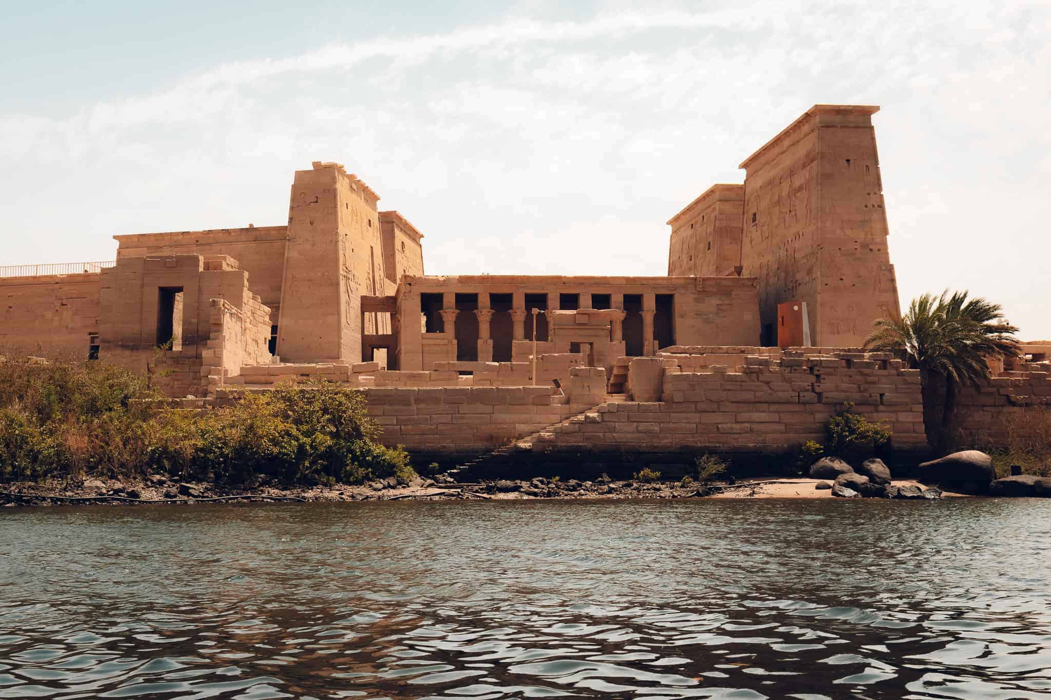 Front view of Philae Temple in Aswan, Egypt, as seen from the Nile River, with its towering pylons, symmetrical columns, and stone steps leading to the water.