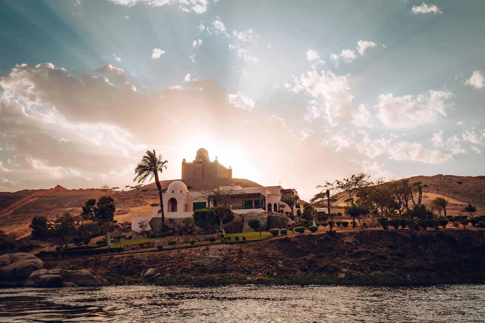 Alt Text:
The Tomb of Aga Khan glows at sunset on a desert hill in Aswan, Egypt, with the domed white mausoleum in the foreground and golden sunrays breaking through clouds.