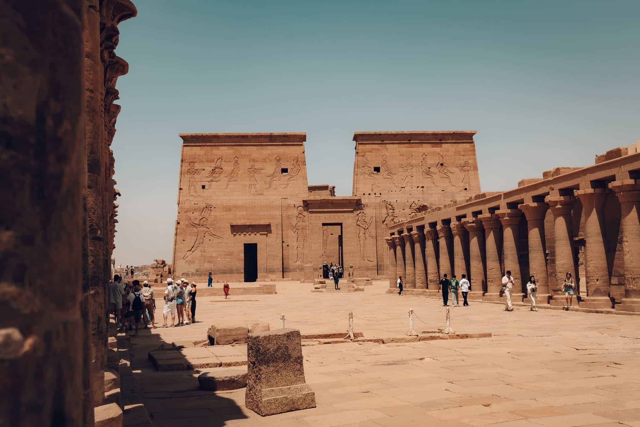Visitors walk across the sunlit courtyard of Philae Temple in Aswan, Egypt, surrounded by tall columned porticos and massive pylons carved with Egyptian deities.