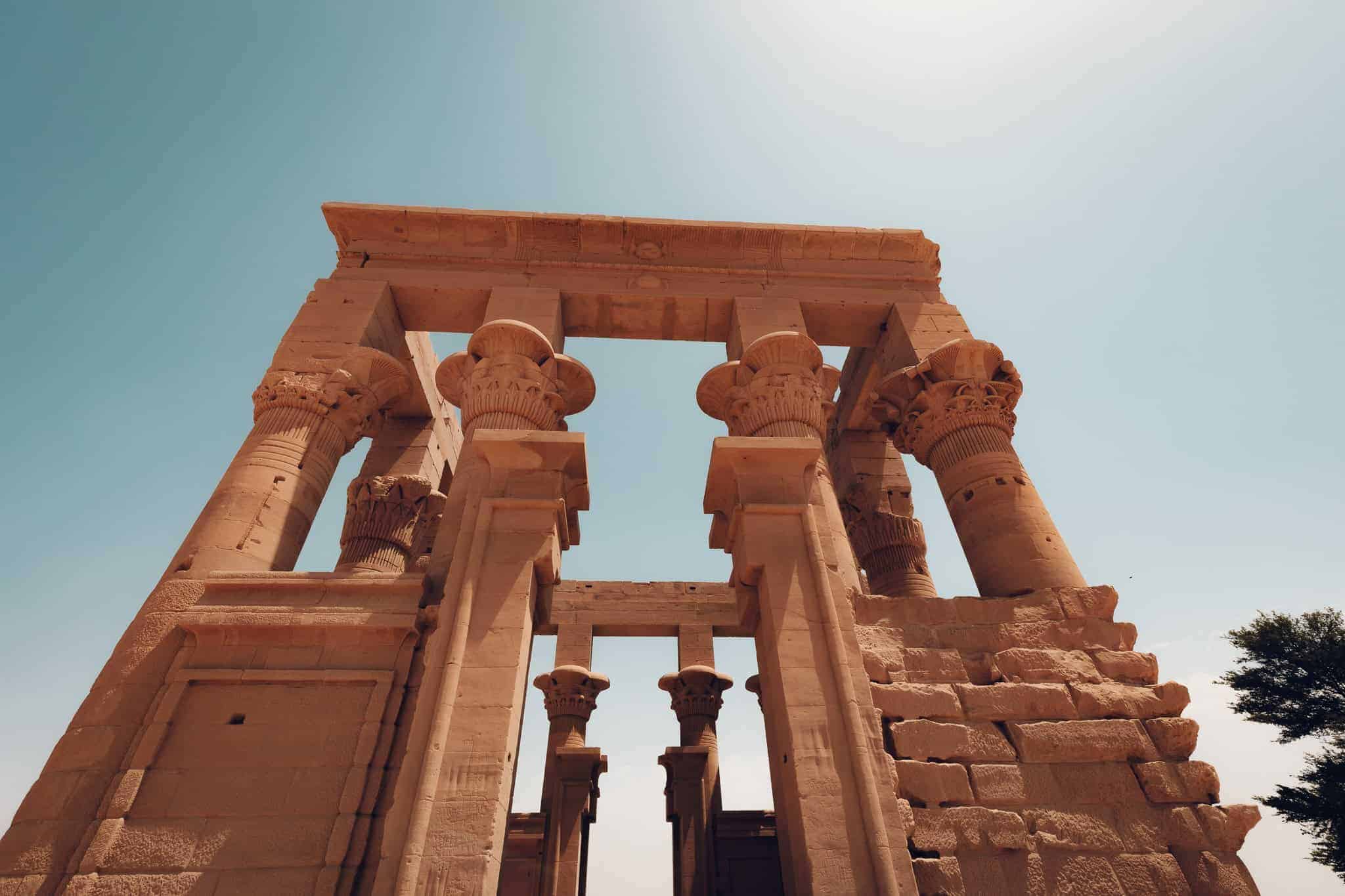 Low angle view of Trajan’s Kiosk at Philae Temple in Aswan, Egypt, with its elegant columns and open roof structure silhouetted against a pale blue sky.