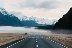 Empty road leading to Milford Sound with snow-capped mountains and rainforest.